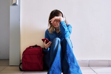 Sad young female student with backpack smartphone sitting on the floor