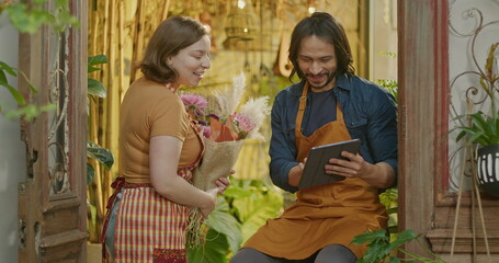 Two employees of flower shop wearing aprons holding tablet and a bouquet of flowers smiling at camera. Male and female workers of a local small business store