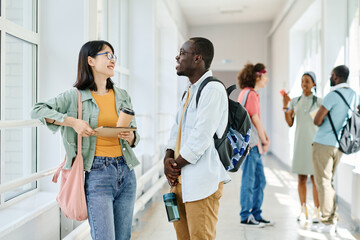 Fototapeta premium Multiethnic students with backpacks chatting during break while standing at school corridor