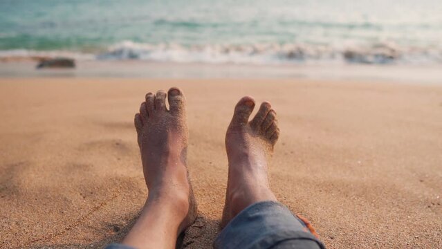 Slow motion POV shot of a man wiggling his sandy toes while relaxing at the beach at Kakolem Beach in Goa, India. Man enjoying his summer holidays at the beach. Summer relaxation background. 
