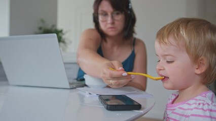 Caucasian Mother Working From Home using Laptop Multitasking Simultaneously Spoon Feeding Baby Girl Daughter Watching Cartoon on Smartphone