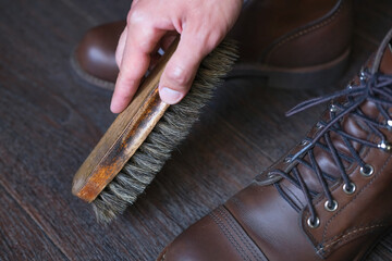 hand with a wooden shoe brush next to stylish leather boot shoes on wooden background