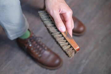 close up of wooden shoe brush in hand against men's shoes boot on wooden floor