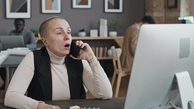 Medium close-up of short-haired Caucasian woman in turtleneck and vest sitting at desk in office at daytime, talking to client on cellphone, choosing paint color