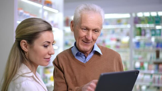Portrait of Caucasian grey-haired senior man talking with pharmacist in drugstore smiling. Positive satisfied client consulting young woman in pharmacy indoors. Service and medicine concept