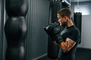 With punching bag. Young tattooed man is practicing boxing indoors in the gym