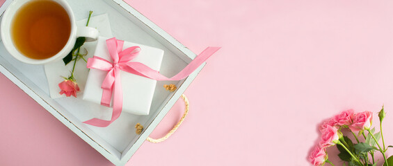 Gift box with tied pink bow and tea in the cup on the white wooden tray on the pink  background.Top view. Copy space.