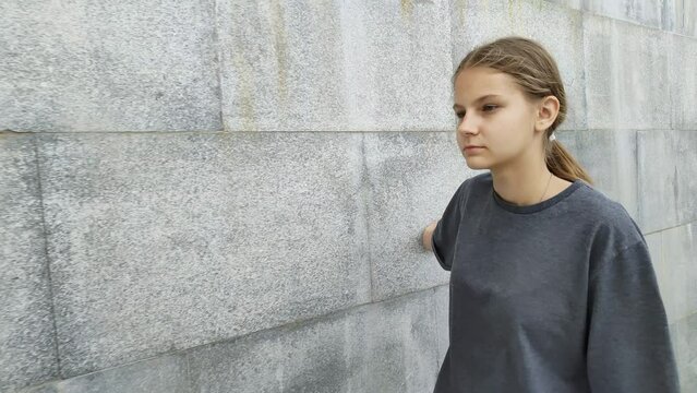 Pretty teenage girl slowly walks along stone wall and touches it with her palm. Sad child walks walks around modern urban area. Thoughtful young woman. Teenage loneliness. Psychology of adolescence