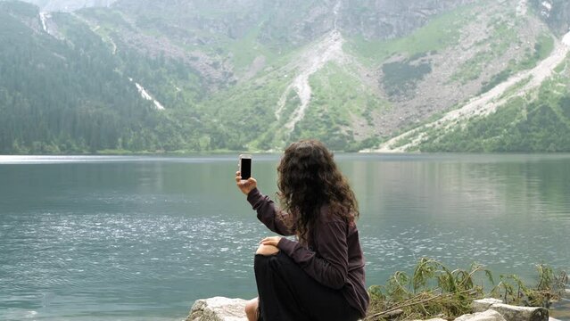 A beautiful girl sits on a rock with her phone in her hands and takes pictures of a snowy mountain.