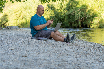 Digital nomad man working with a laptop and a phone, sitting on the bank of a river. 