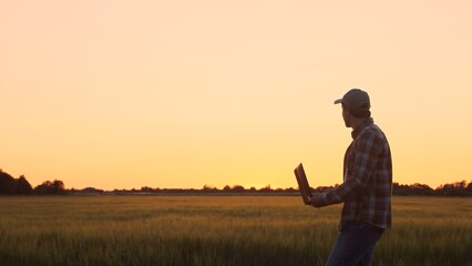 Farmer with a laptop computer in front of a sunset agricultural landscape. Man in a countryside field. Country life, food production, farming and technology concept.