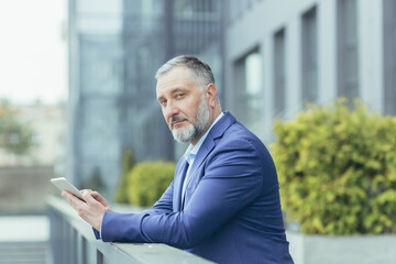 Portrait of serious and concentrated senior and experienced broker investor, gray-haired male outside office looking thoughtfully at camera, businessman in business suit holding tablet computer