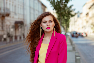 Fashionable brunette woman with long natural hair, pink fuchsia color lips makeup, wearing trendy earrings, classic blazer, walking in street of European city. Copy, empty space for text