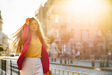 Fashionable woman with long hair wearing trendy yellow sunglasses, pink fuchsia blazer yellow blouse, walking in street of European city. Copy, empty space for text