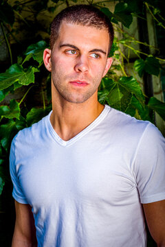 Young man with short hair, wearing white V neck T shirt, standing outdoors by green ivy leavers in New York City, looking away.