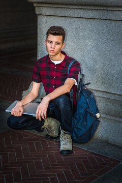 Young man wearing short sleeve, black, red patterned shirt, jeans, boot shoes, carrying shoulder bag, holding laptop computer,  squatting on corner of street in New York City, sad, relaxing, thinking.