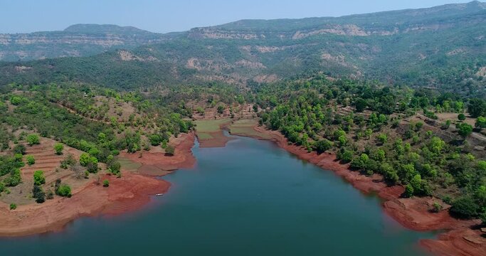 Aerial shot of river running along forest with reddish soil, hill background