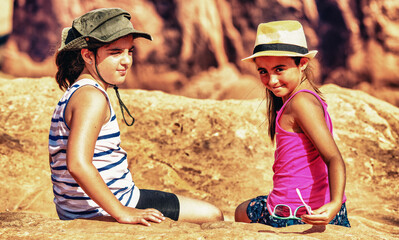 Two young girl wearing straw hats seated on a national park rock under the sun.