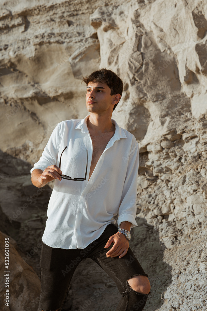 Sticker Portrait of handsome sexy young man in white shirt posing staying on sand stones on the beach near sea holding sunglasses in his hand.