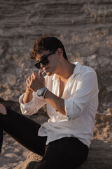 Portrait of handsome sexy young man in white shirt siting on sand stones on the beach near sea at sunset, smoking, lighting a cigarette..