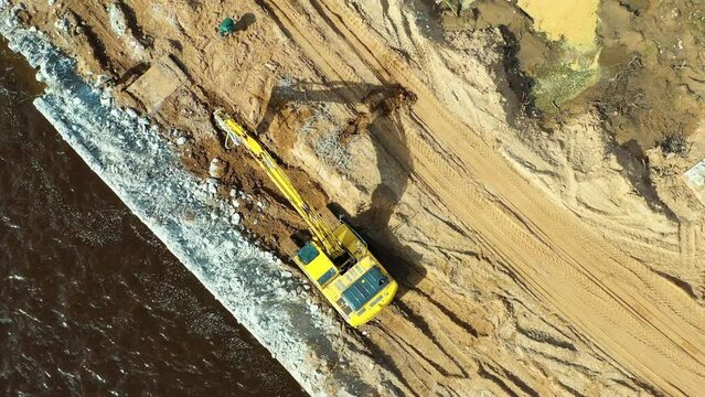 An excavator with a jackhammer works on the banks of the river, the construction of the embankment, aerial view