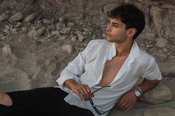 Portrait of handsome sexy young man in white shirt holding sunglasses in hand sitting on the sand...