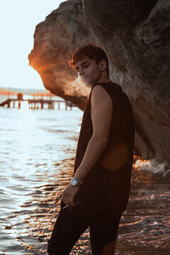 Handsome sexy young man in black t-shirt  staying near sand stones on the beach near sea at sunset, smoling cigarette.