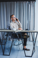 Indoor shot of cheerful bearded male freelancer work at distant, manages finances via smart phone, sits at wooden table, works with documentation, has happy expression. People, work, technology