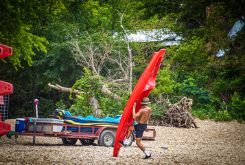 Man with no shirt struggling to load red plastic kayak on float trip trailer on pebbly river beach...