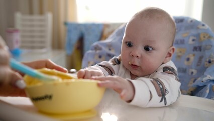 Young mother feeding her baby son with fruit puree. Cute baby eating solid food from a spoon. Mom...