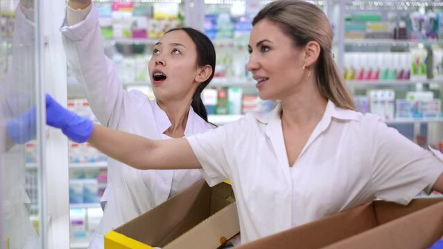Portrait of positive Asian new employee talking with professional Caucasian pharmacist in drugstore putting pills on shelves. Beautiful young women chatting working in pharmacy restocking