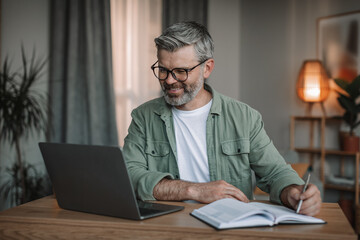 Glad senior european male with beard in glasses watches video on computer, makes notes at table in room