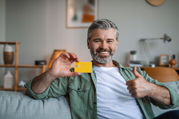 Satisfied retired european man with beard shows credit card for shopping online and thumb up, hand...