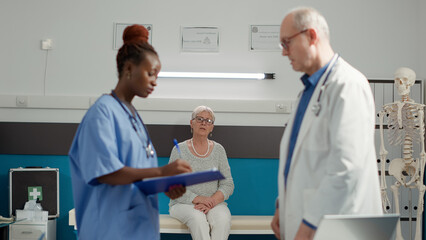 Fototapeta premium Retired woman waiting to receive sickness diagnosis from medical staff in cabinet, to cure disease with medicine. Sitting in doctors office to find exam results from diverse team of specialists.