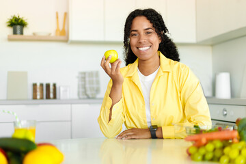 Obraz premium Weight loss diet. Happy black lady holding apple and smiling at camera, sitting in modern kitchen interior, copy space