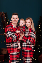 Obraz na płótnie Canvas Christmas Family holidays. Smiling young dad, mom are hugging baby boy son in the same red checked pyjamas near the decorations Christmas tree with light at home. Happiness. New Years concept