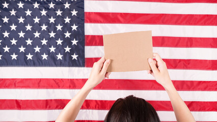 Woman is holding an empty cardboard with Space for Text sign US flag on background. Protest against...