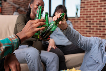 Multiethnic group of friends clinking bottles of beer at home, saying cheers and making toast to celebrate reunion. Happy people toasting with alcoholic drinks, celebrating event.