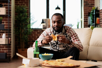 Relaxed person eating slice of pizza in front of television at home, having takeaway delivery meal...