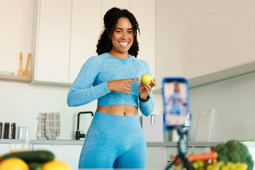 Healthy food blog. Happy black fit lady recording video for her food blog on smartphone, standing in kitchen - obrazy, fototapety, plakaty