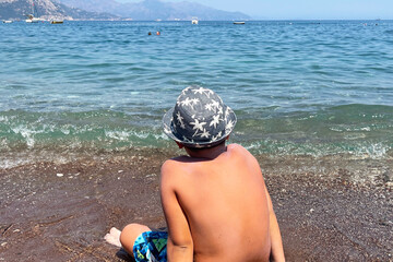 The boy in the hat is looking towards the sea. Back-to-back child watching the sea on the beach during summer vacation. Kid is sitting on the sand on the beach.