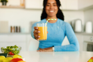 Happy black fitness lady in sportswear showing fresh orange juice in glass, sitting in kitchen after domestic workout,