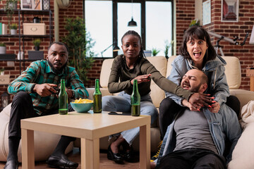 Happy group of friends having fun with video games strategy, playing competition on tv console and drinking beer. Young people enjoying house party together, leisure activity.
