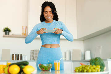 Food and fitness blogging concept. Fit black lady taking photo of healthy dinner on smartphone, standing in kitchen
