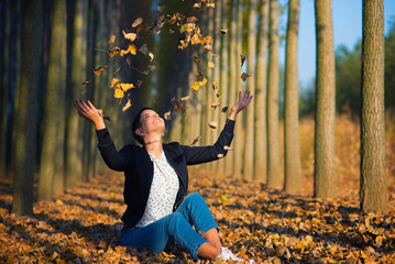 Beautiful smiling girl throws falling dry forest leaves in the air on a sunny autumn day