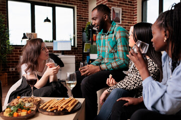 Happy friends discussing at wine party while celebrating birthday event. Multiracial group of happy young people enjoying alcoholic beverage while telling childhood stories.