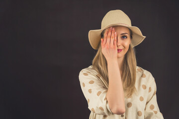 Fototapeta premium Young white blonde woman wearing linen polka-dot shirt and straw hat covering one eye with hand looking into camera. Dark background studio shot. High quality photo