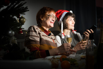 Cheerful mature woman celebrating New Year at home with her adult daughter, watching festive tv...