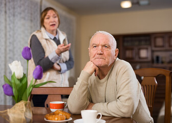 Offended elderly man sitting at home table on background with angry wife reprimanding him