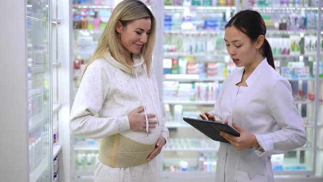 Pregnant woman putting on maternity belt talking with pharmacist in drugstore. Beautiful Caucasian and Asian women choosing belly belt in pharmacy standing with digital tablet